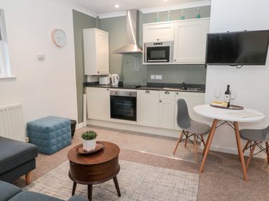 A kitchen area with white cabinets and appliances next to a small dining table with two chairs and a living area with a round wooden coffee table and a blue ottoman at Flodden in Crookham near Cornhill-On-Tweed