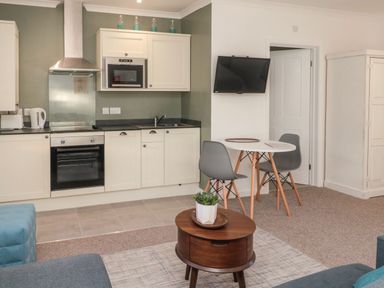 A kitchen area with white cabinets and appliances next to a small dining table with two chairs and a wall-mounted TV at Flodden in Crookham near Cornhill-On-Tweed