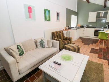 A living room with a beige sofa and leaf print cushions a striped armchair with a footstool a white coffee table and a kitchen area at Bakers Cottage in Crookham near Cornhill-On-Tweed