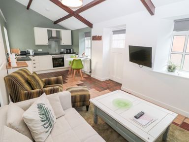 A living area with a beige sofa a striped armchair and ottoman a white coffee table a wall-mounted TV a small dining table with green chairs and a kitchen with white cabinets and appliances at Bakers Cottage in Crookham near Cornhill-On-Tweed