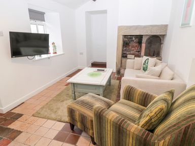 A living room with a wall-mounted TV a striped armchair a beige sofa a white coffee table and a stone fireplace at Bakers Cottage in Crookham near Cornhill-On-Tweed