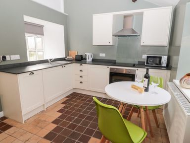 A kitchen with white cabinets black countertops a stove microwave and a round table with green chairs at Bakers Cottage in Crookham near Cornhill-On-Tweed