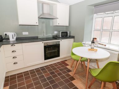 A kitchen with white cabinets a black countertop green chairs around a white table and a window at Bakers Cottage in Crookham near Cornhill-On-Tweed