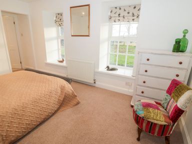 A bedroom with a bed covered in a beige blanket two windows with bird patterned blinds a white chest of drawers with green glass bottles and a colorful patchwork chair at Bakers Cottage in Crookham near Cornhill-On-Tweed
