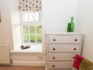 A window with a duck figurine on the sill next to a white chest of drawers with green glass bottles and a multicolored cushion at Bakers Cottage in Crookham near Cornhill On Tweed