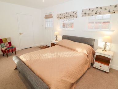 A bedroom with a double bed covered with a beige quilt two bedside tables with lamps a door two windows with patterned blinds and a multicolored chair Bakers Cottage Crookham near Cornhill-On-Tweed