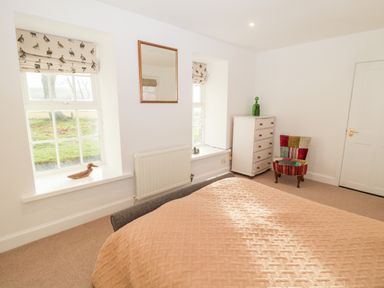 A bedroom with two windows featuring bird patterned blinds a bed with a quilt a white chest of drawers a patchwork upholstered chair and a wall mirror at Bakers Cottage in Crookham near Cornhill-On-Tweed