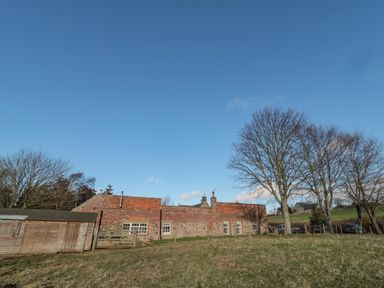 A brick and stone building with multiple windows near a wooden shed and leafless trees on grassy land at Bakers Cottage in Crookham near Cornhill-On-Tweed
