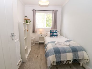 A small bedroom with a single bed a bedside table with a lamp a window with curtains a shelving unit and light wood flooring at Valley Pines in Valley