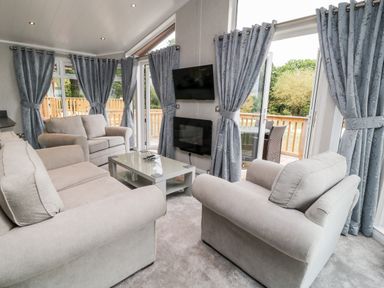 A living room with beige sofas a glass coffee table wall mounted tv and windows with blue curtains at Woodpecker Lodge in Highampton near Sheepwash