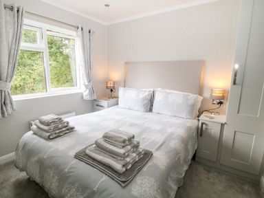 A bedroom with a double bed two bedside tables lamps folded towels on the bed and a window with curtains at Woodpecker Lodge Highampton near Sheepwash
