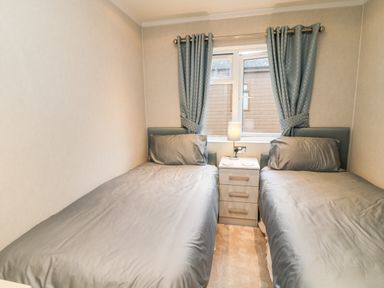 A small bedroom with two single beds separated by a nightstand with a lamp and blue curtains on the window at Woodpecker Lodge in Highampton near Sheepwash