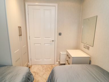 A bedroom with two beds a white door a white wardrobe a white chest of drawers with a mirror above it and a radiator at Woodpecker Lodge in Highampton near Sheepwash