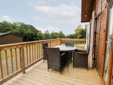 An outdoor wooden deck with a round glass table and four wicker chairs at Woodpecker Lodge in Highampton near Sheepwash