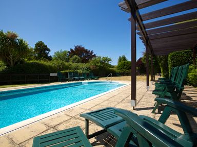 An outdoor swimming pool with green lounge chairs and a pergola surrounded by trees and bushes at 14 St Elmo Court in Salcombe