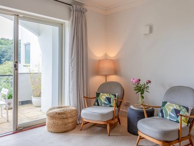 A sitting area with two chairs with cushions a lamp a side table with flowers and a woven ottoman near sliding glass doors at 14 St Elmo Court in Salcombe