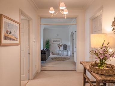 A hallway with a wooden table holding a lamp and flowers and an open view to a living room with a dark sofa and side tables at 14 St Elmo Court in Salcombe