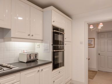 A kitchen with white cabinets a double oven a toaster and a gas stove next to a doorway showing a hallway with a framed picture and chandelier at 14 St Elmo Court in Salcombe