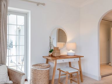 A wooden dressing table with a round mirror and lamp beside a woven basket and stool near a glass door at 14 St Elmo Court in Salcombe