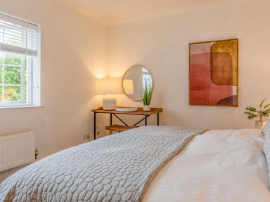 A bedroom with a bed covered by a blanket a window a wooden desk with a lamp a round mirror and a wall painting at 14 St Elmo Court in Salcombe