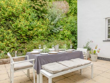 An outdoor dining area with a table covered with a cloth set with plates and glasses surrounded by white chairs and a bench with cushions at 14 St Elmo Court in Salcombe