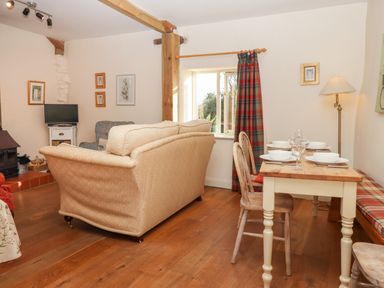 A living room with a dining table set with dishes and glasses a beige sofa wooden chairs a window with plaid curtains and a small television at The Old Dairy in Colyton