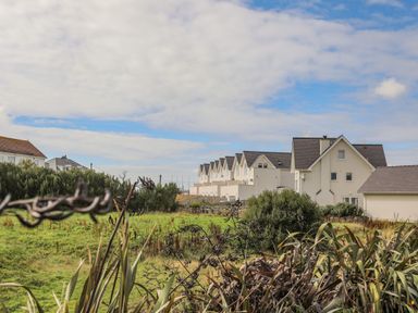 A row of white houses with pitched roofs behind green shrubs and grass at 9 Lon Y Dryw in Trearddur Bay