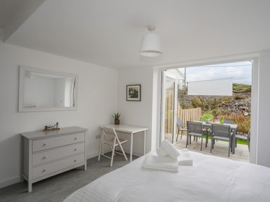 A bedroom with a bed and folded towels a table with a chair a dresser and an open door leading to an outdoor seating area at 9 Lon Y Dryw in Trearddur Bay