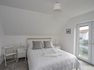 A bedroom with a bed and pillows bedside tables lamps a folding chair and a door with a window at 9 Lon Y Dryw in Trearddur Bay