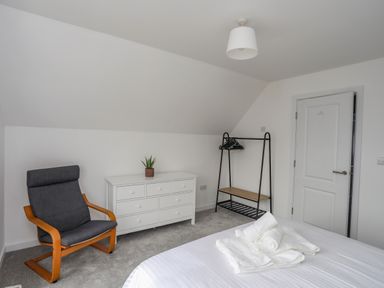 A bedroom with a bed with folded towels a white chest of drawers a black clothes rack and a gray chair at 9 Lon Y Dryw in Trearddur Bay