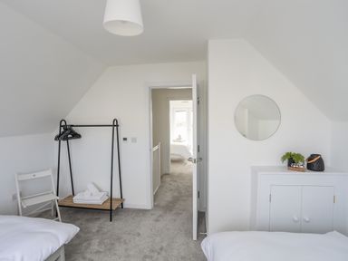 A bedroom with beds a clothing rack a chair a cabinet with decoration and an open door leading to another room at 9 Lon Y Dryw in Trearddur Bay