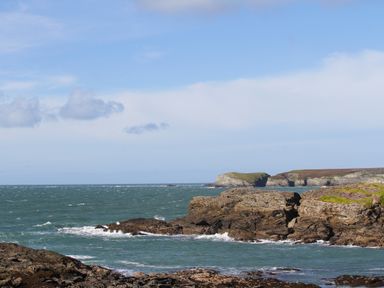 A rocky coastline with waves crashing against the shore and cliffs in the distance at 9 Lon Y Dryw in Trearddur Bay