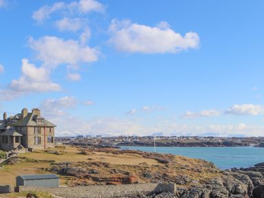 A stone house near a rocky coastline with water and distant houses under a blue sky at 9 Lon Y Dryw in Trearddur Bay