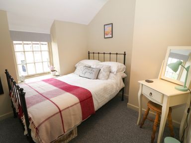 A bedroom with a bed covered in white and red blankets next to a window and a white vanity table with a green lamp and wooden stool at Llety Bach in Caernarfon