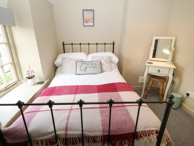 A bedroom with a metal bed frame a red and white blanket a small white dressing table with a mirror and lamp and a window with a pink flower at Llety Bach in Caernarfon