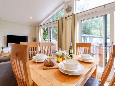 A dining area with a wooden table set with white dishes and bowls glasses bread and fruit next to large windows with curtains at Garuda Calgarth 19 White Cross Bay near Windermere