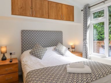 A bedroom with a double bed with patterned headboard and cushions two bedside tables with lamps and wooden cabinets above the bed at Garuda Calgarth 19 White Cross Bay near Windermere