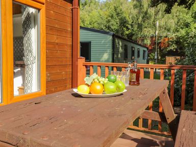 A wooden outdoor table with a plate of fruit and bottles on a balcony at Garuda Calgarth 19 White Cross Bay near Windermere