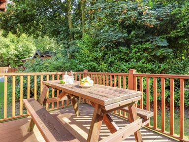 An outdoor wooden picnic table with a bowl of fruit and glass bottles on the table on a deck with wooden railing surrounded by green trees and bushes at Garuda Calgarth 19 White Cross Bay near Windermere