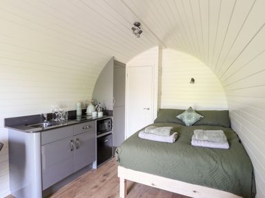 A small room with a bed with green bedding and folded towels a kitchenette with a sink glasses a kettle and microwave at Tyddyn Parthle Pod 1 in Bontnewydd Gwynedd