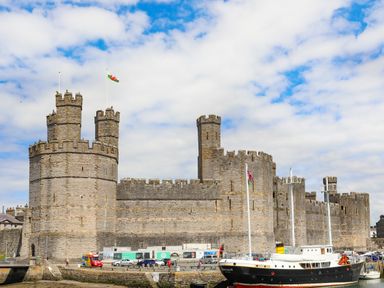 A stone castle with multiple towers near a dock with boats and parked cars at Tyddyn Parthle Pod 1 in Bontnewydd Gwynedd