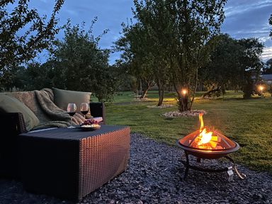 An outdoor seating area with a wicker sofa and table with wine glasses and snacks next to a fire pit in a garden at Tyddyn Parthle Pod 1 in Bontnewydd Gwynedd