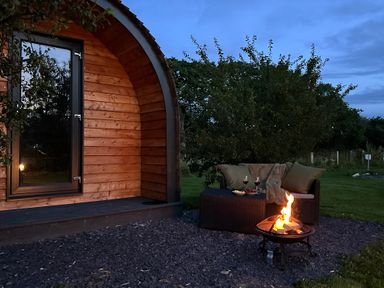 A wooden pod with a glass door next to a fire pit and an outdoor sofa with cushions and blankets at Tyddyn Parthle Pod 1 in Bontnewydd Gwynedd