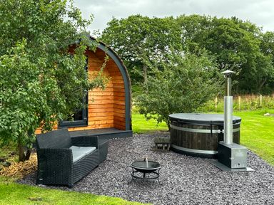 An outdoor area with a wooden pod building a black wicker sofa a fire pit and a covered hot tub in grass at Tyddyn Parthle Pod 1 Bontnewydd Gwynedd