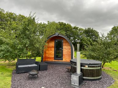 A small wooden cabin with a glass door surrounded by green trees and a lawn with outdoor furniture and a hot tub at Tyddyn Parthle Pod 1 in Bontnewydd Gwynedd