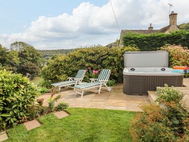 An outdoor patio with two striped lounge chairs a hot tub and surrounding greenery at The Gables in Sheepscombe