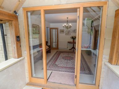 An entrance with glass wooden doors opening into a room with a patterned rug a chandelier wall pictures a wooden bench and a round table with flowers at The Gables in Sheepscombe