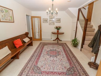 A hallway with a wooden bench and cushions a patterned rug a small table with flowers a staircase and framed pictures at The Gables in Sheepscombe