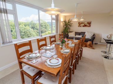 A dining room with a wooden table set for six people near large windows a brown sofa with cushions and a dog on the floor at The Gables in Sheepscombe