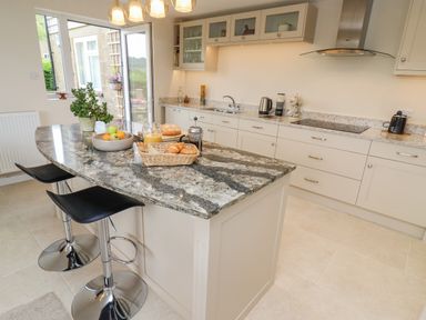 A kitchen with a granite island countertop with fruit bowl and breakfast items two black stools and cream cabinets at The Gables in Sheepscombe